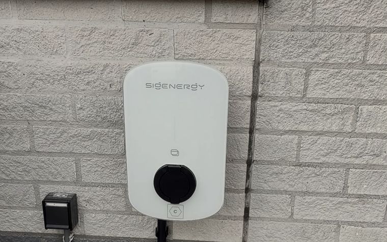 Laadpaal aan de buitenmuur van een woning, met de aanvoerkabel in een zwarte goot.